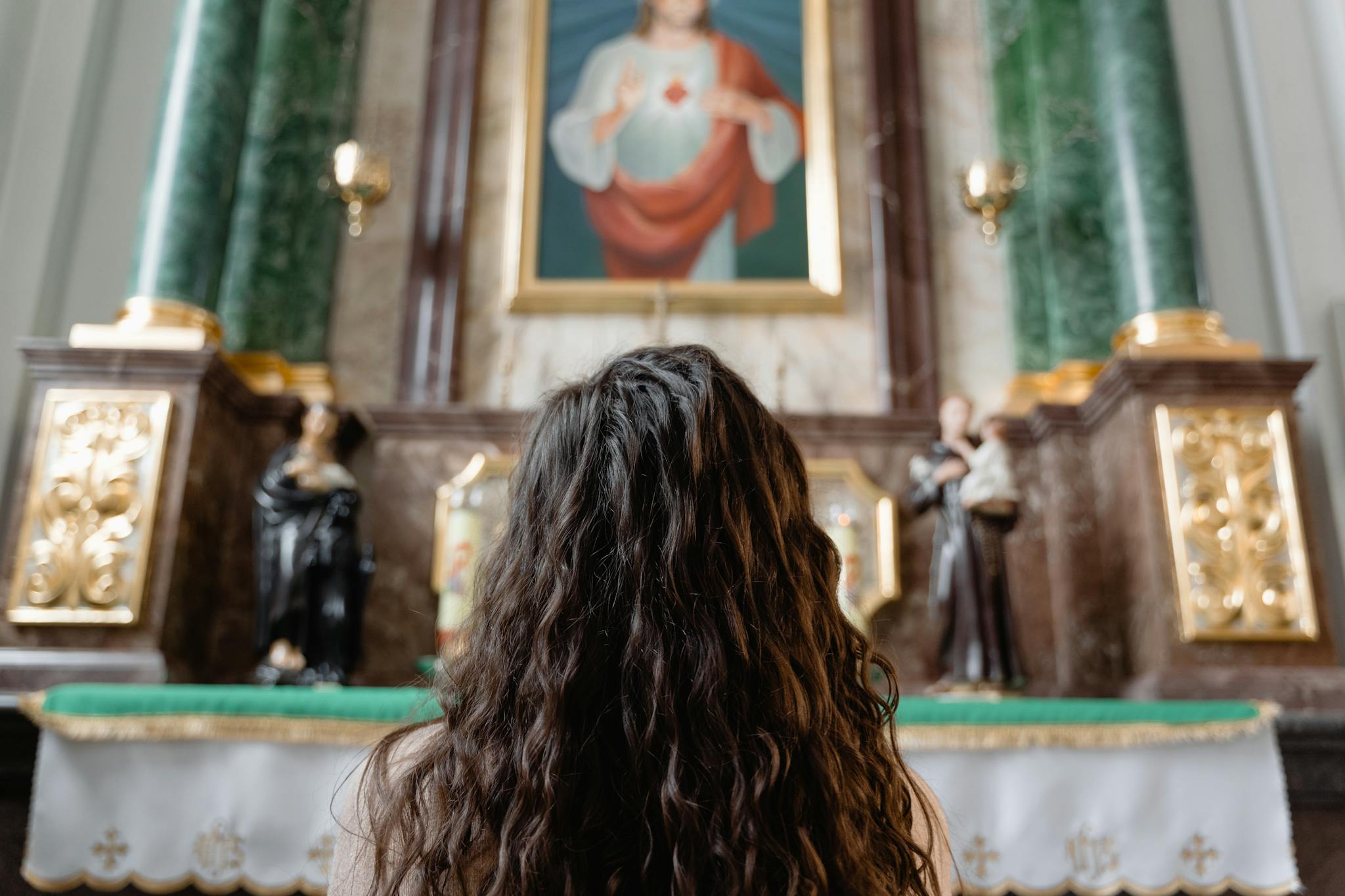 Woman with curly hair kneeling in prayer before an ornate church altar featuring religious icons.